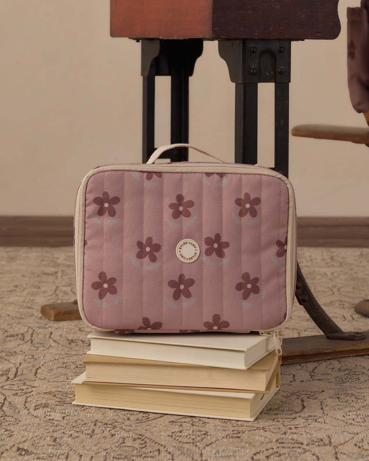 A rylee + cru lunch bag in little daisy rests atop a stack of four closed books on a patterned carpet, next to dark metal furniture legs.