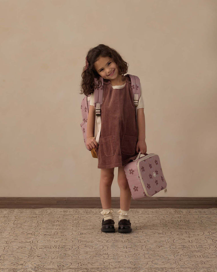 A young girl with curly hair smiles, wearing a pink dress and white shirt, holding the rylee + cru lunch bag in little daisy and wearing a matching star-patterned backpack, standing against a beige background.