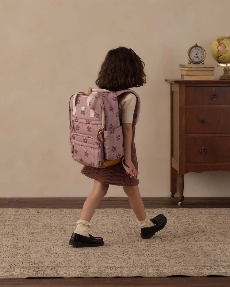 A young girl with short brown hair, wearing a rylee + cru backpack in little daisy, a white shirt, brown skirt, and black shoes, walks past a wooden dresser topped with books, a clock, and a globe—ready for her everyday adventures.
