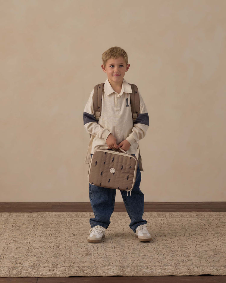 A young child stands on a patterned rug against a neutral beige wall, wearing a cream long-sleeve shirt, blue jeans, white sneakers, and a backpack, holding the rylee + cru lunch bag in bolts—a stylish insulated lunch bag in beige quilted fabric.