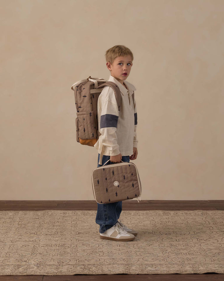 A young boy stands on a patterned rug against a beige background, dressed in jeans, a white shirt, and white sneakers. He carries a tan backpack along with the rylee + cru lunch bag in bolts—an insulated and stylish bag featuring small dark prints.