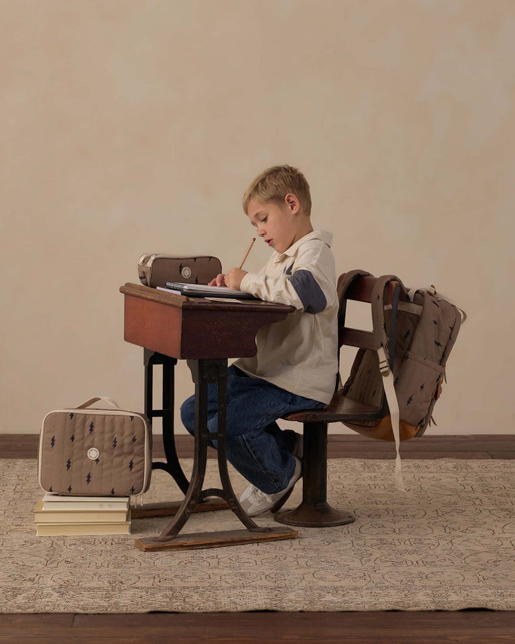 A young boy sits at a vintage wooden desk, writing in a notebook. He is surrounded by school supplies and bags, with the rylee + cru lunch bag in bolts resting stylishly beside his books on the floor, a backpack hanging on the chair, and more books stacked nearby.