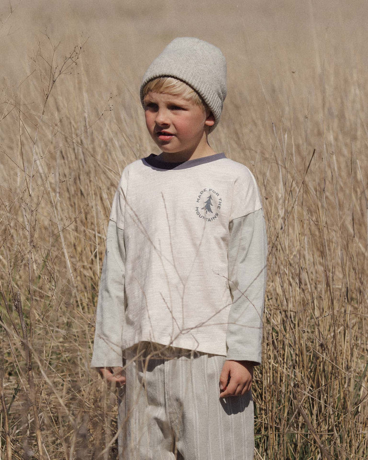 A young child wearing a gray beanie and the rylee + cru PREORDER camden long sleeve tee in made for the mountains, crafted from 100% cotton, stands in a dry, grassy field and gazes slightly to the side under soft natural light.