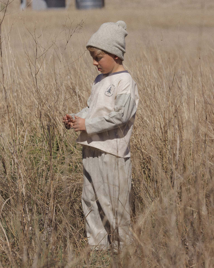 A young child wearing a gray beanie and a light-colored PREORDER Camden Long Sleeve Tee in Made for the Mountains by rylee + cru stands in a field of tall, dry grass, looking down at something in their hands.