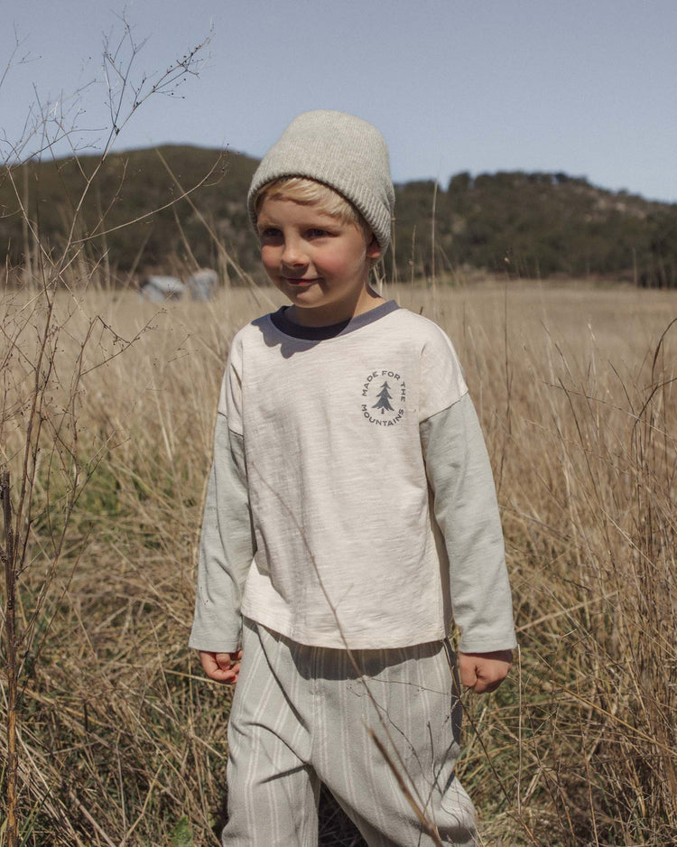 A young child wearing a light-colored beanie, striped pants, and the rylee + cru PREORDER camden long sleeve tee in made for the mountains stands in a dry, grassy field with hills and trees in the background under a clear sky.