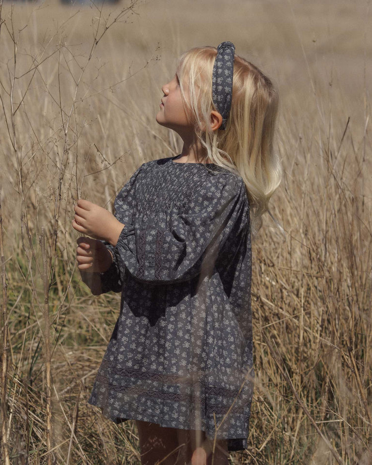 A young child with long blonde hair, wearing a gray floral dress and the rylee + cru knotted headband in heart ditsy, stands in a field of dry tall grass, looking up and touching a stem.