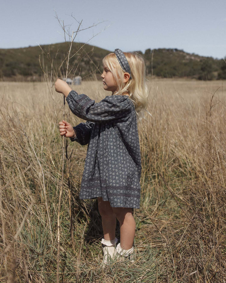 A young girl wearing the selene dress in heart ditsy by rylee + cru, featuring puff sleeves and a subtle heart ditsy print, stands in a grassy field holding a tall dry plant. She completes the look with a matching headband. Hills and trees stretch out in the background beneath a clear sky.