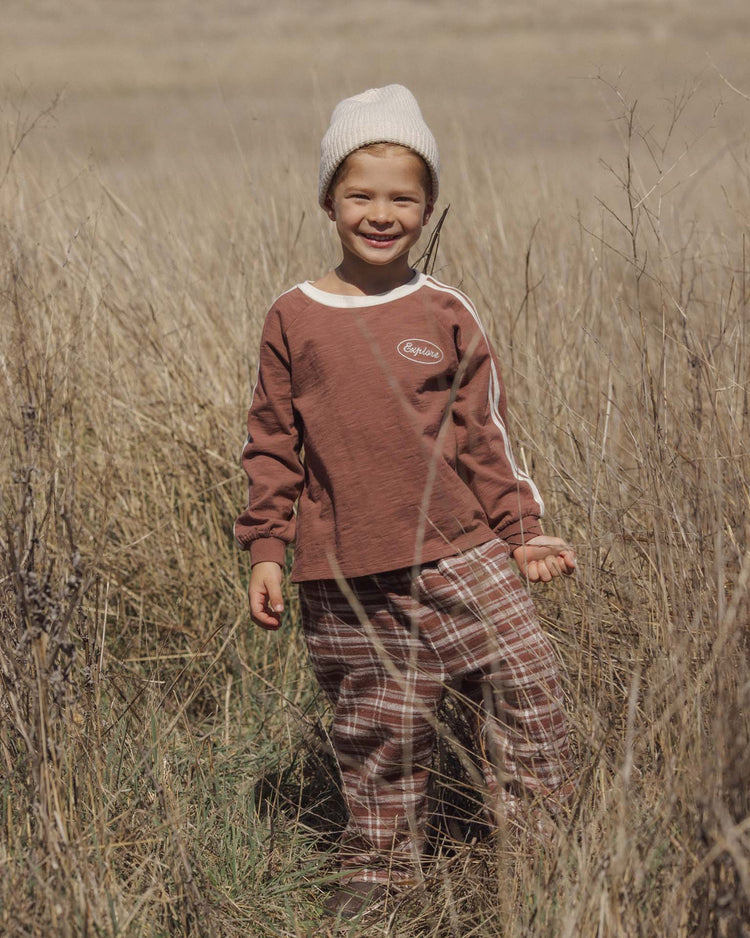 A smiling child in a knit hat, brown long-sleeve shirt, and rylee + cru palmer pant in aspen plaid with an elastic waistband and baggy fit stands in a dry grassy field on a sunny day.