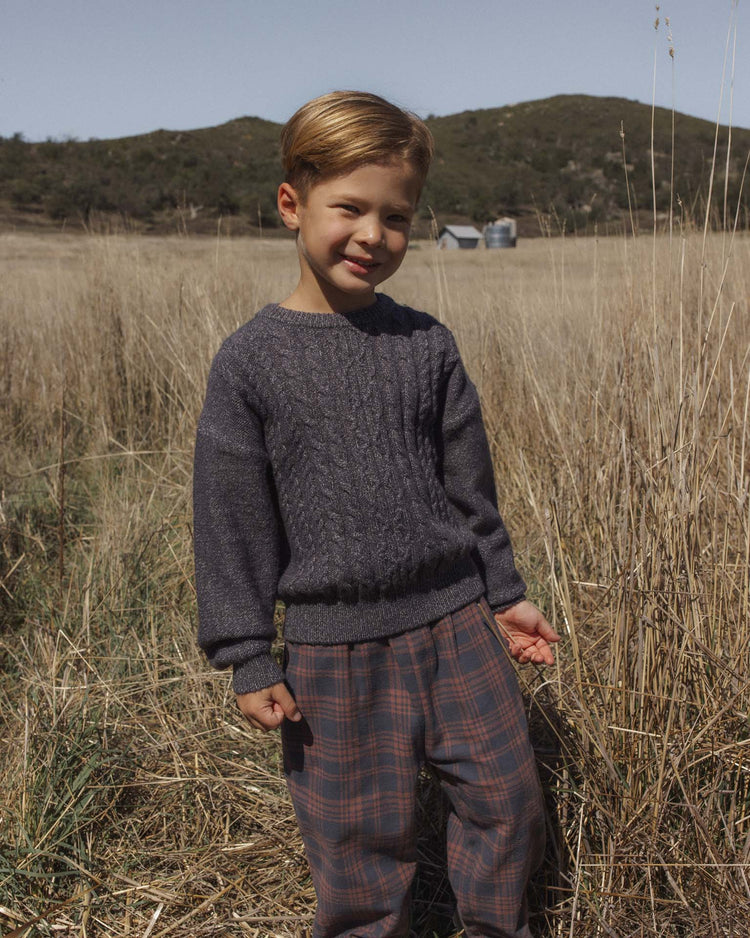 A young child with light brown hair, dressed in a gray sweater and rylee + cru’s palmer pant in midnight plaid with a baggy fit, stands smiling amid a dry grassy field, hills, and a few small buildings under a clear sky.
