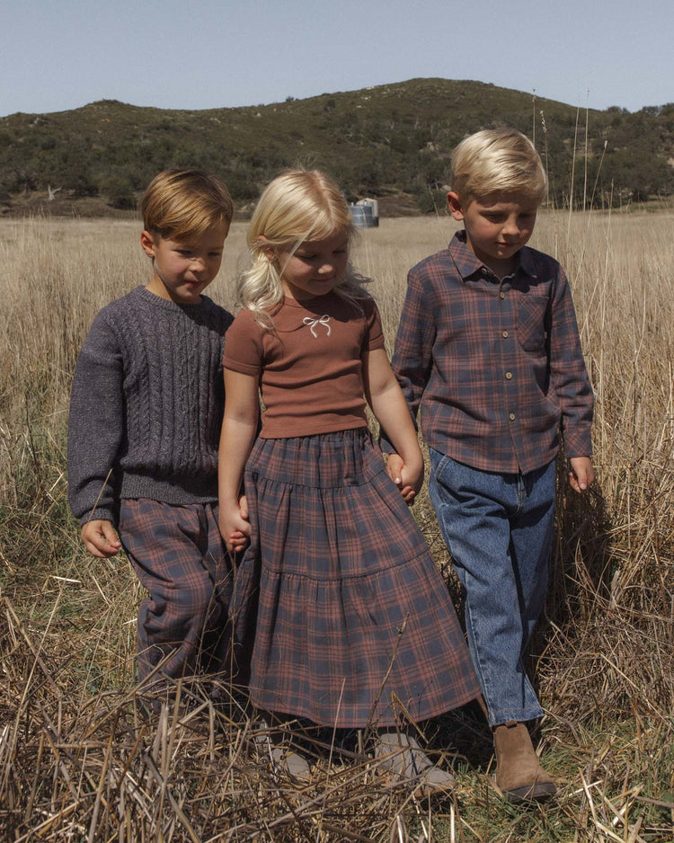 Three young children walk hand in hand through a grassy field, dressed for fall in earthy tones. Each wears cozy rylee + cru palmer pant in midnight plaid, featuring a comfortable baggy fit. Rolling hills stretch beneath a clear sky behind them.