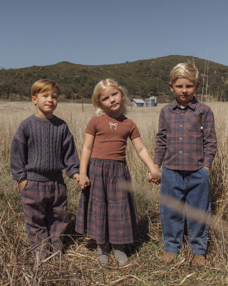 Three young children stand in a dry grassy field, holding hands. Two boys wear rylee + cru collared long sleeve shirts in midnight plaid with pants; the girl in the middle wears a brown top and plaid skirt. Hills and a barn are visible in the background under a clear sky.