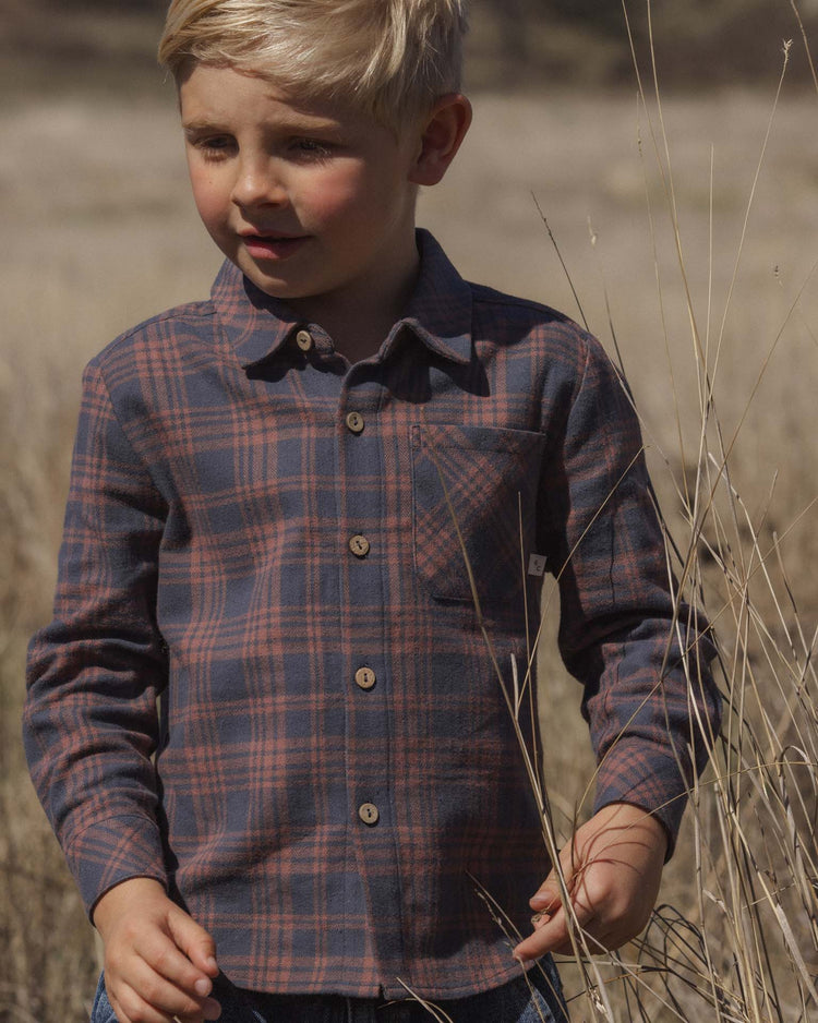 A young child with blonde hair wearing the rylee + cru collared long sleeve shirt in midnight plaid stands outside in a dry, grassy field, looking to the side.