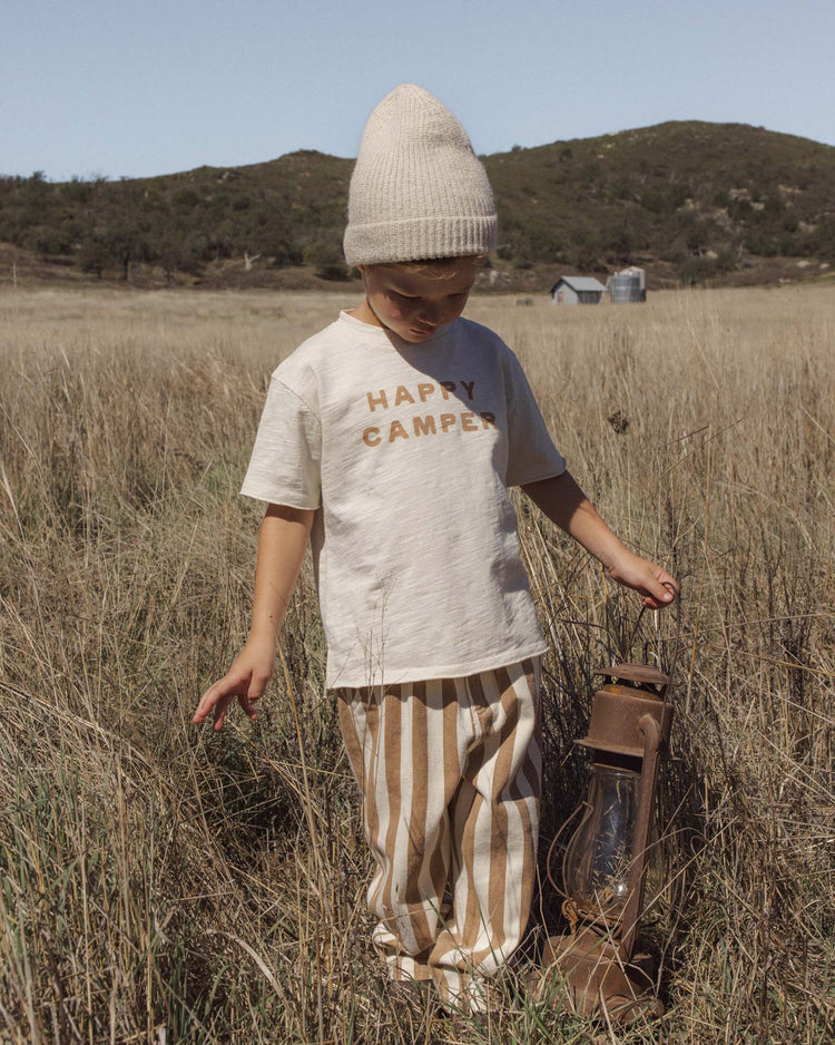 A child wearing rylee + cru’s baggy pant in fawn pinstripe, a Happy Camper shirt, and a knit hat stands in a grassy field holding a rustic lantern, with hills and a barn in the background.