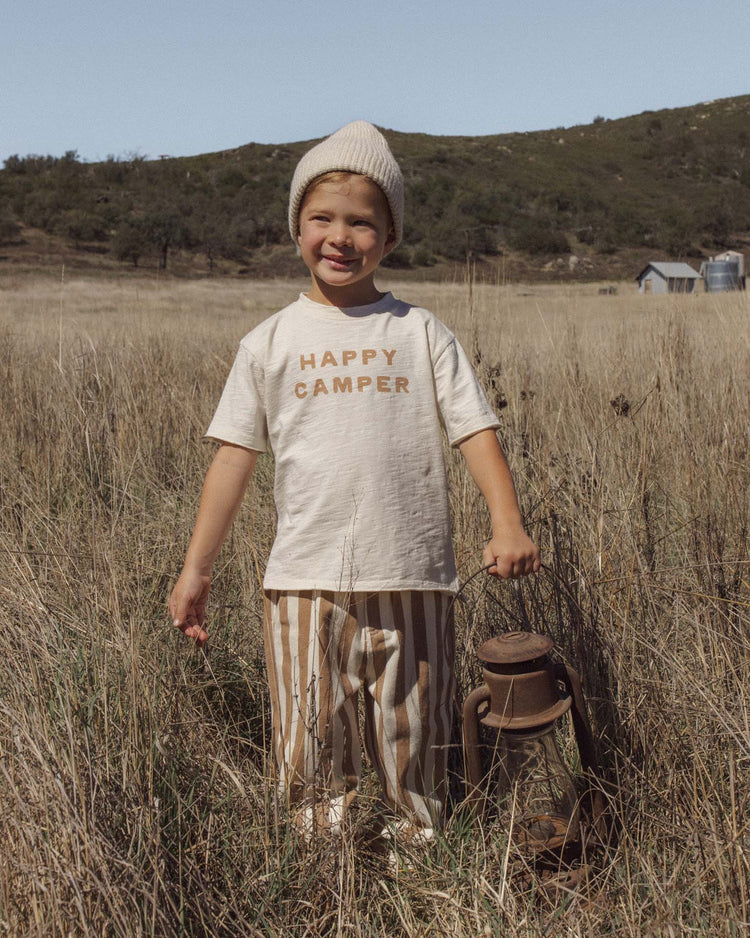 A child wearing rylee + cru’s baggy pant in fawn pinstripe with an elastic waistband and a HAPPY CAMPER shirt stands in a grassy field, holding a lantern and smiling. Hills, trees, and a small building are visible in the background.