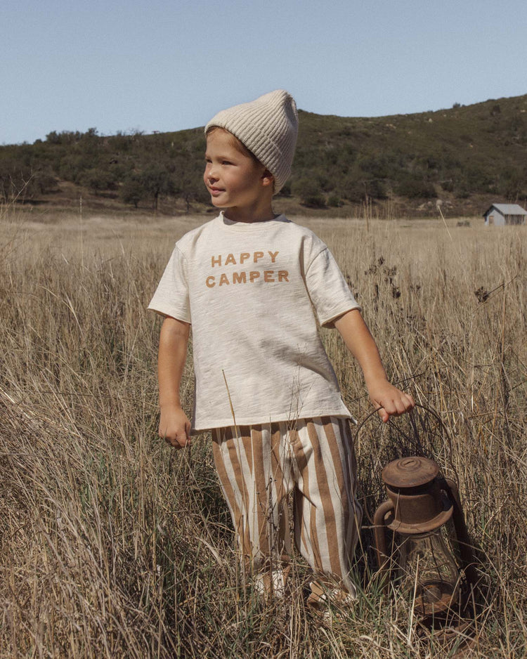 A young child in a HAPPY CAMPER shirt, rylee + cru’s baggy pant in fawn pinstripe with an elastic waistband, and a knit hat stands in a grassy field holding a lantern. Hills and a small building are visible in the background.
