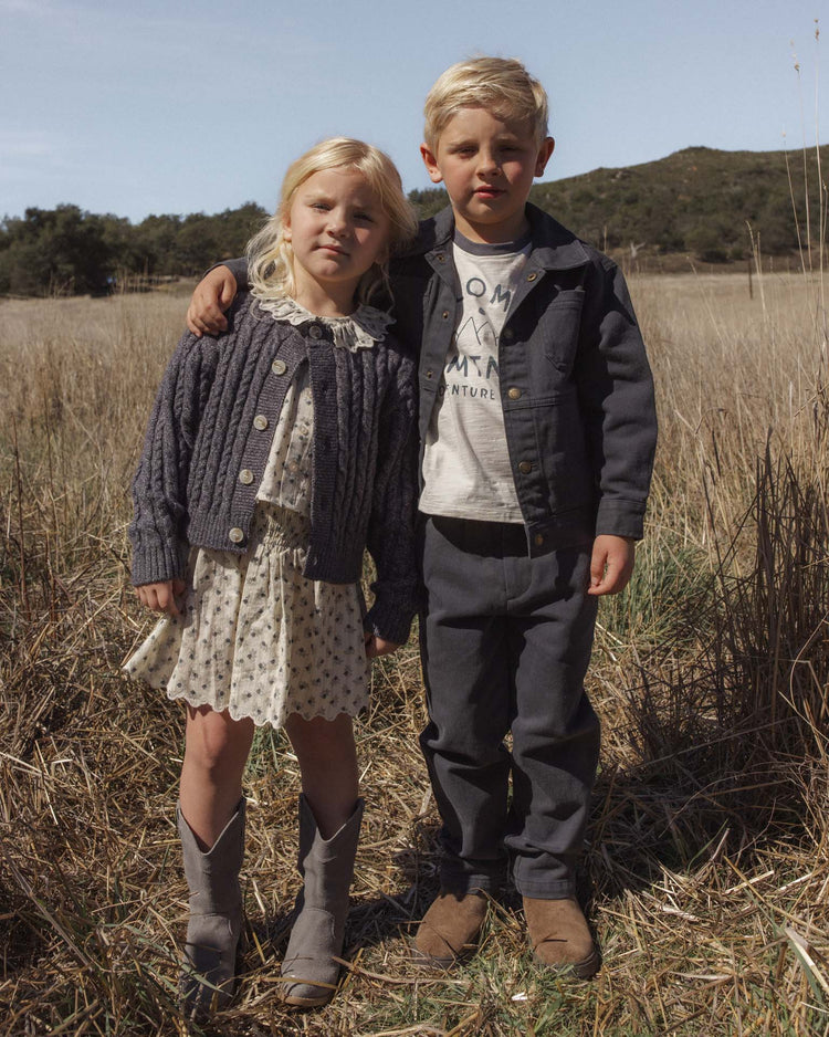 Two young children stand side by side in a grassy field, the boy’s arm around the girl’s shoulder. Both have blond hair and neutral expressions, dressed in casual autumn clothing—the girl wears the rylee + cru cheyenne cardigan in midnight, featuring distinctive marble buttons that add a cozy layer to her look. Trees and hills can be seen in the background.