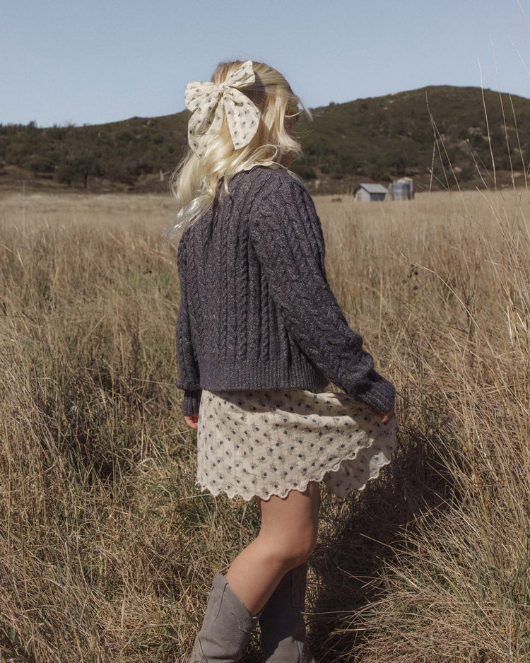 A person with long blonde hair, wearing a large polka-dot bow, the rylee + cru cheyenne cardigan in midnight featuring marble buttons, a patterned dress, and boots, stands in a field of tall grass facing away. Hills and a barn are visible in the distance beneath a clear sky.