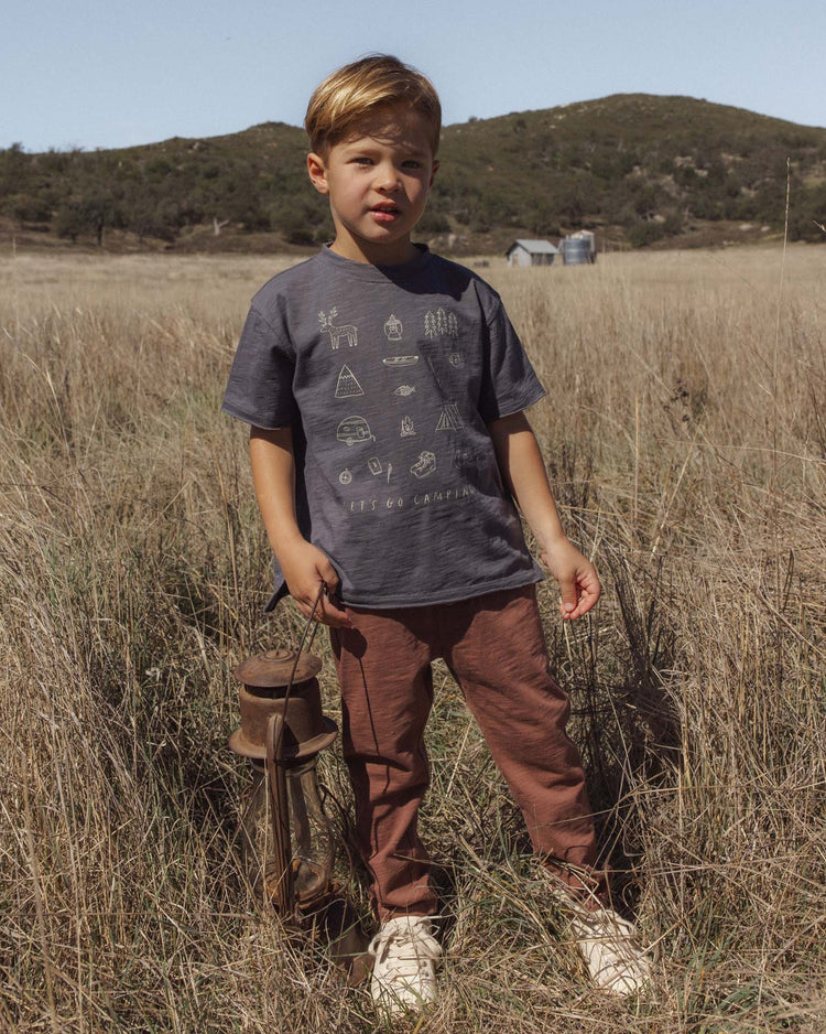 A young boy stands in a dry grassy field holding a lantern, wearing a dark t-shirt with outdoor symbols, PREORDER rylee + cru cru pant in mulberry, and white shoes. Hills and a small building are visible in the background under a clear sky.
