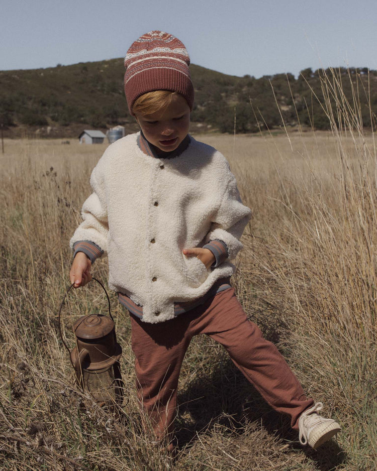 A child wearing the rylee + cru PREORDER cru pant in mulberry stands in a grassy field, paired with a white jacket and a knit hat, holding a lantern with hills and a house in the background.