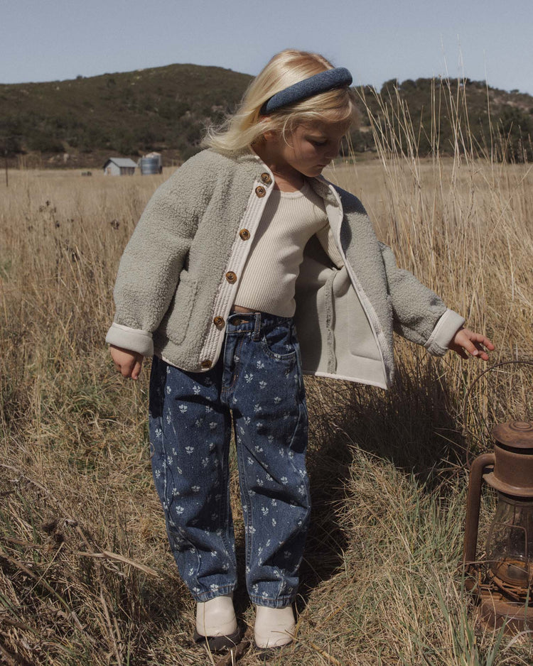 A young child with blonde hair stands in a grassy field wearing a gray jacket, indigo 100% cotton denim jeans, and the rylee + cru padded headband in indigo, looking down at an old lantern on the ground.