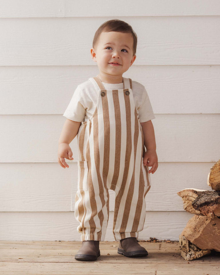 A toddler dressed in a white shirt and the rylee + cru cargo baby overall in fawn pinstripe—crafted from 100% cotton—stands indoors on a wooden floor, smiling softly, with stacked firewood seen to the right.