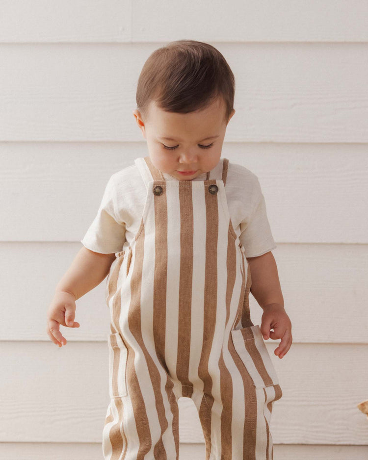 A toddler wearing a cream shirt and the rylee + cru cargo baby overall in fawn pinstripe, made from 100% cotton, stands in front of a white wooden wall, looking down with hands slightly raised.