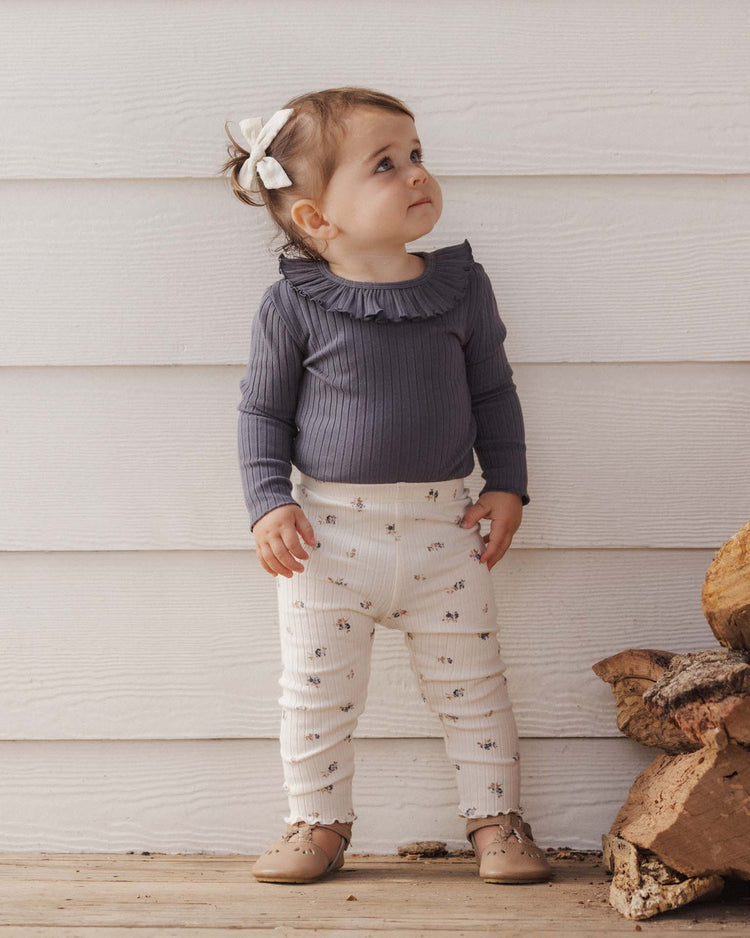 A toddler with brown hair and a white bow, wearing a purple long-sleeve shirt and the PREORDER legging in natural ditsy by rylee + cru, stands indoors on a wooden floor next to a small stack of firewood, looking up.