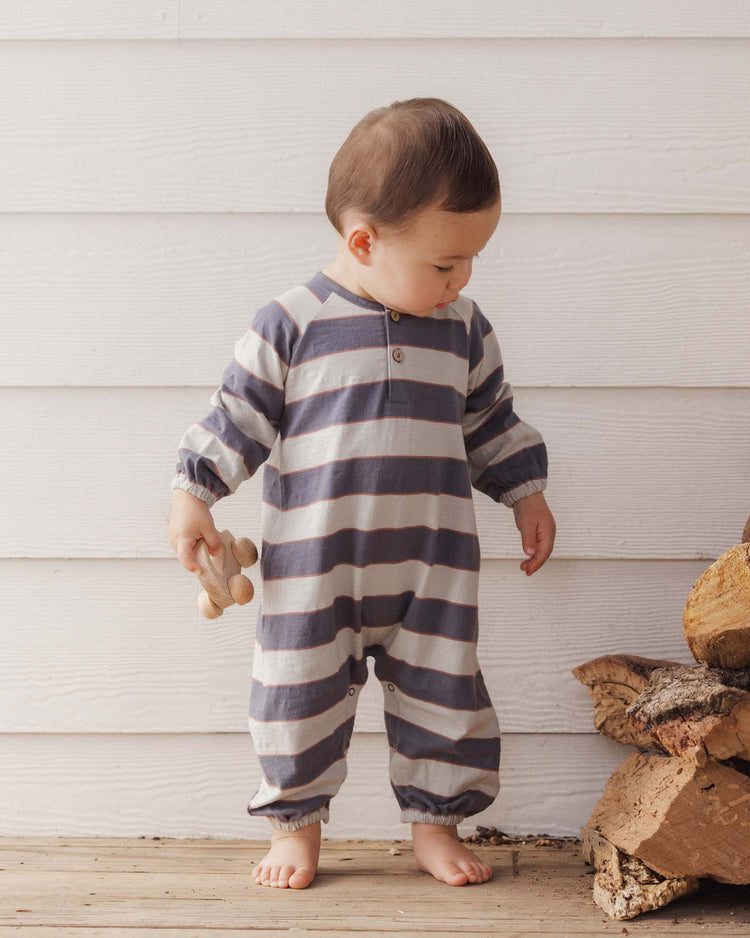 A toddler wearing the rylee + cru henley jumpsuit in sage stripe, crafted from 100% cotton, stands barefoot on a wooden floor. The child is holding a wooden toy and looking down, with a stack of firewood to the right and a white wooden wall in the background.