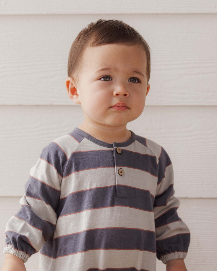 A young child with short brown hair, wearing the rylee + cru henley jumpsuit in sage stripe (100% cotton), stands in front of a white wooden wall and gazes slightly to the side with a neutral expression.