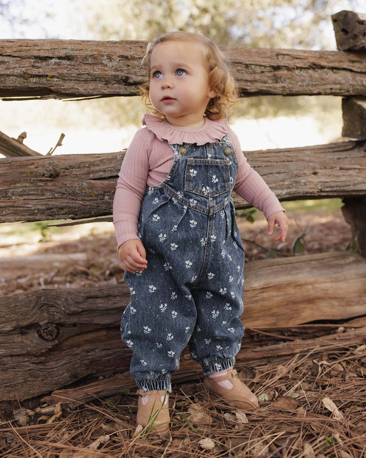 A toddler with curly blonde hair, dressed in a pink long-sleeve top and the rylee + cru vintage overall in heart ditsy—crafted from 100% cotton—stands outdoors by a rustic wooden fence on a bed of pine needles.
