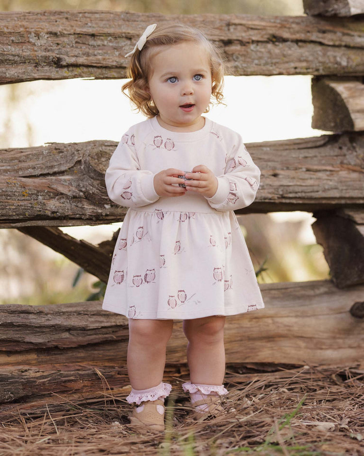 A young girl with curly hair stands outdoors in front of a wooden fence, looking up with a curious expression, wearing the rylee + cru PREORDER raglan dress in owls.
