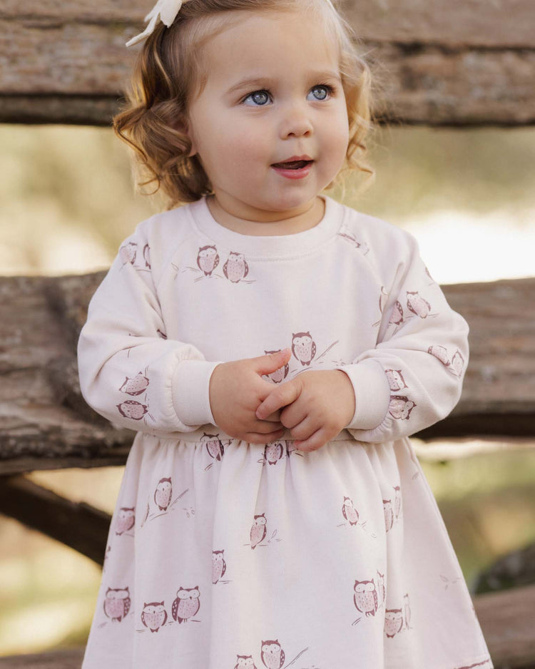 A young girl with curly blonde hair stands outdoors in front of rustic wooden beams, looking up with a slight smile, wearing the rylee + cru PREORDER raglan dress in owls.