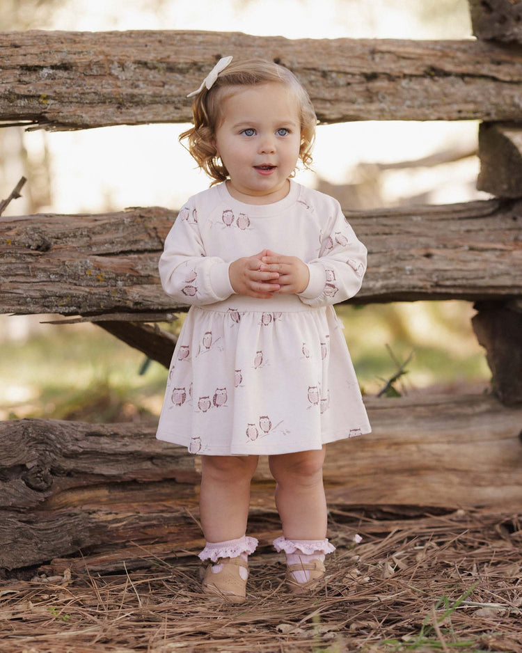 A young child with light brown hair stands outdoors in front of a rustic wooden fence, gazing slightly to the side while wearing the rylee + cru PREORDER raglan dress in owls and matching socks.