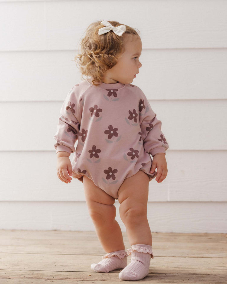 A toddler with curly hair, wearing a bow, stands on a wooden floor facing sideways in front of a white wall, dressed in the rylee + cru PREORDER crew neck romper in daisy.