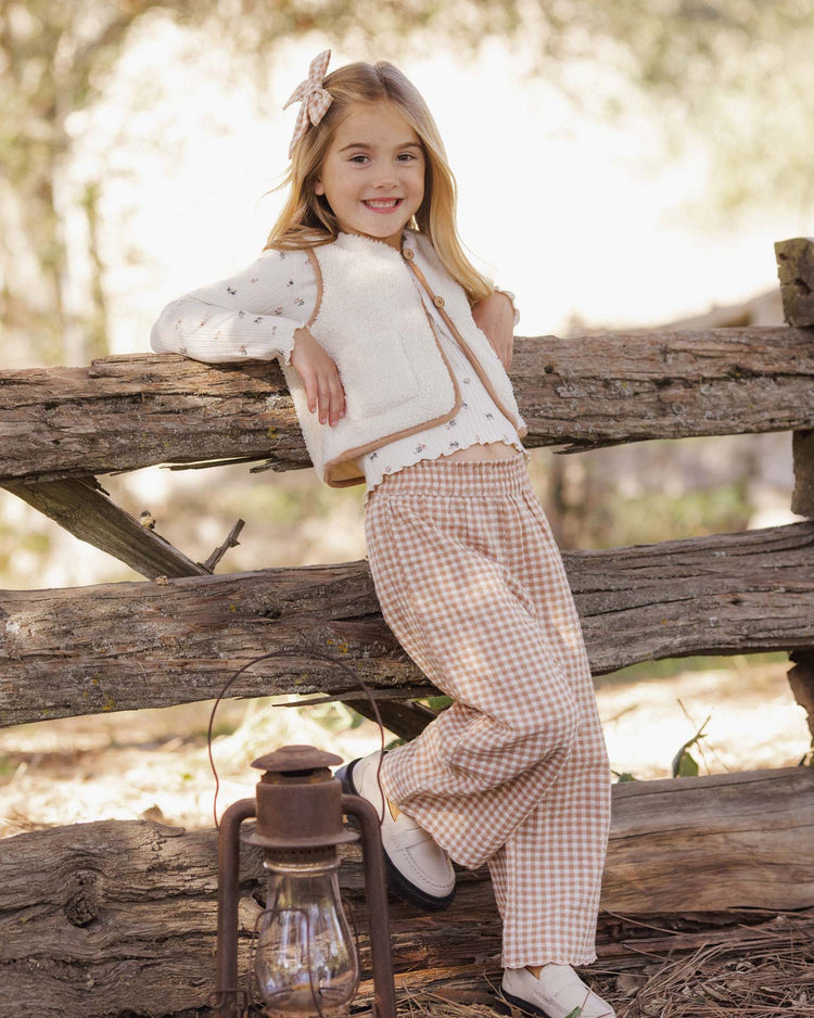 A young girl in checkered pants and a cozy vest smiles as she leans against a wooden fence outdoors. A bow adorns her hair, and a lantern rests on the ground nearby. Her rylee + cru ribbed long sleeve in natural ditsy, featuring a lace-trimmed neckline, adds a charming touch to the bright and rustic scene.