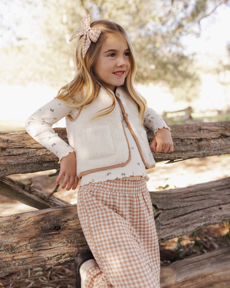 A young girl with long blonde hair, wearing the rylee + cru ribbed long sleeve in natural ditsy beneath a cream vest, paired with a floral top, gingham pants, and a matching bow, leans against a wooden fence outdoors surrounded by trees. Her outfit features delicate lace-trimmed details and charming hints of natural ditsy print.