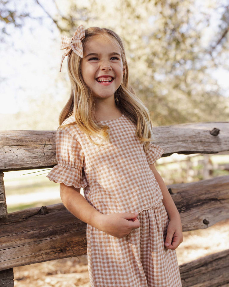 A smiling young girl stands by a rustic wooden fence outdoors, bathed in soft sunlight filtering through the trees, wearing the rylee + cru francie blouse in fawn gingham with a cropped fit and matching bow.