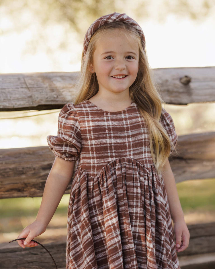 A young girl with long blonde hair is smiling outdoors in front of a wooden fence, wearing a brown and white dress paired with the rylee + cru padded headband in aspen plaid, made from 100% cotton; blurred trees can be seen in the background.