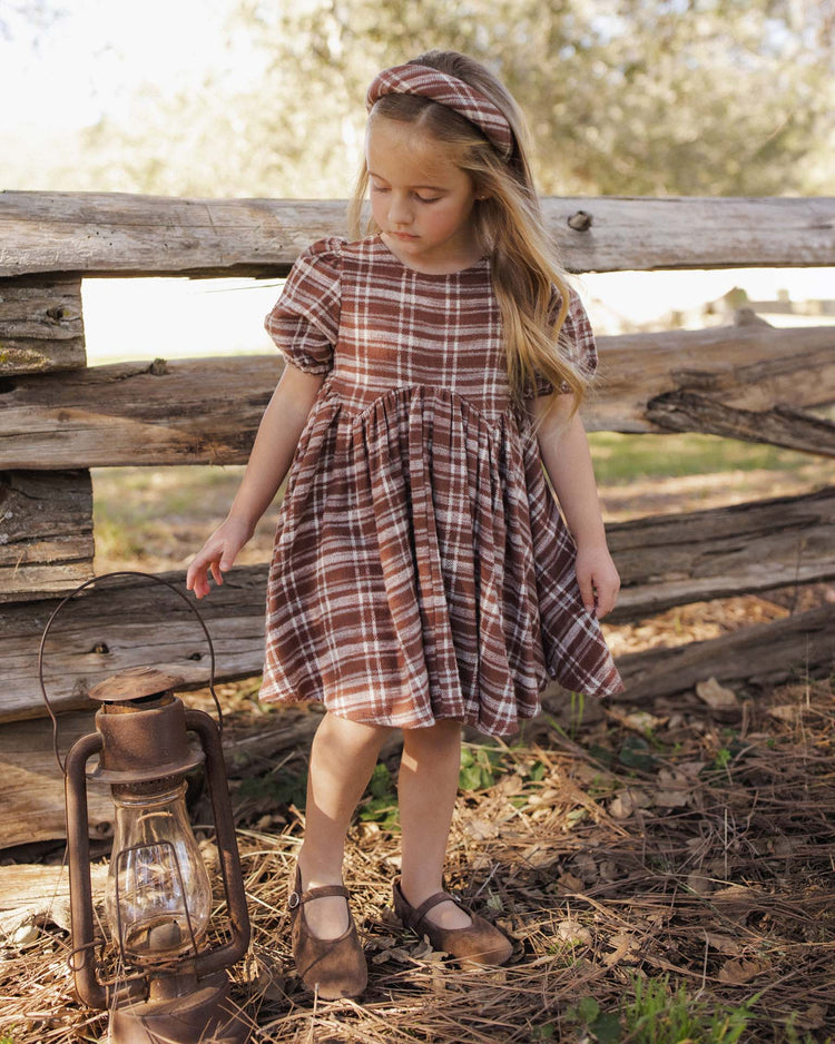 A young girl wearing the rylee + cru padded headband in aspen plaid stands by a wooden fence, holding a vintage lantern. Sunlight and greenery frame the scene, while her 100% cotton outfit blends comfort with timeless style.