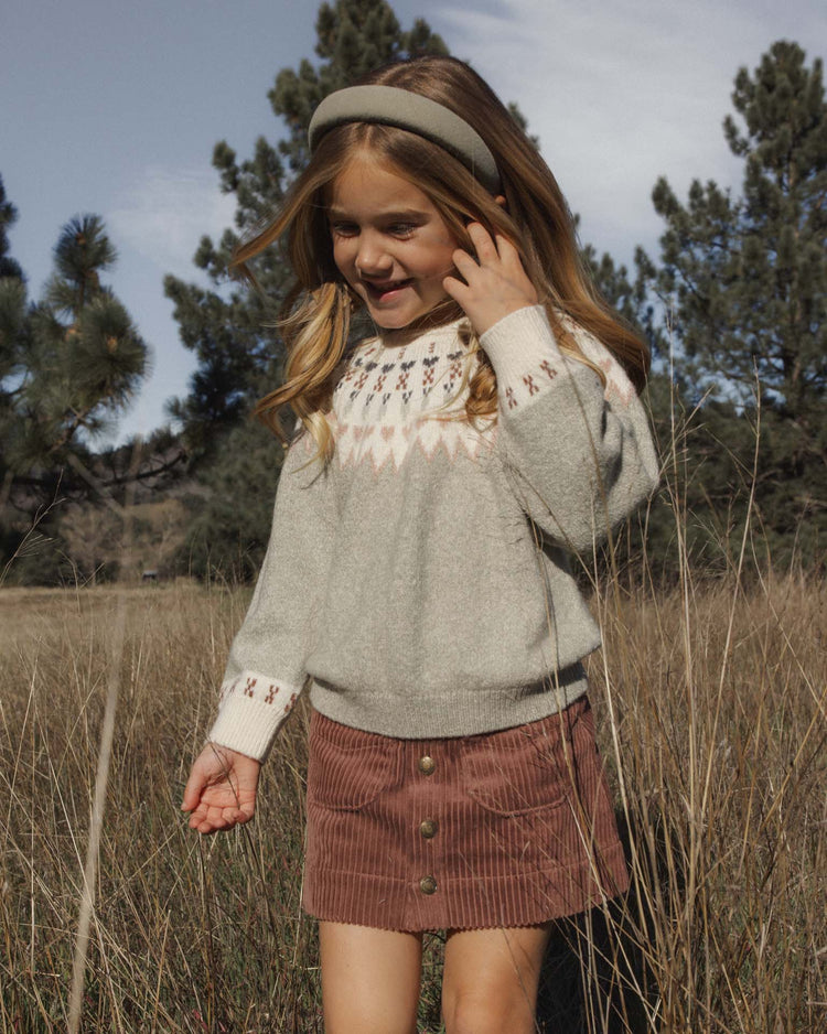 A young girl stands in a grassy field, wearing a patterned sweater, a dark sage corduroy skirt, and the rylee + cru padded headband in dark sage. Trees and a partly cloudy sky are in the background.