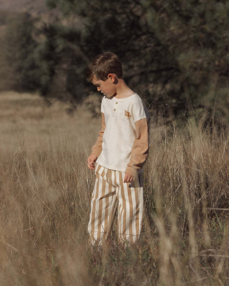 A young boy with brown hair stands in tall dry grass, wearing a cream shirt with tan sleeves and the rylee + cru baggy pant in fawn pinstripe, featuring an elastic waistband. He looks down, surrounded by trees and blurred nature in the background. This relaxed outfit is crafted from 100% cotton, combining comfort and natural style.