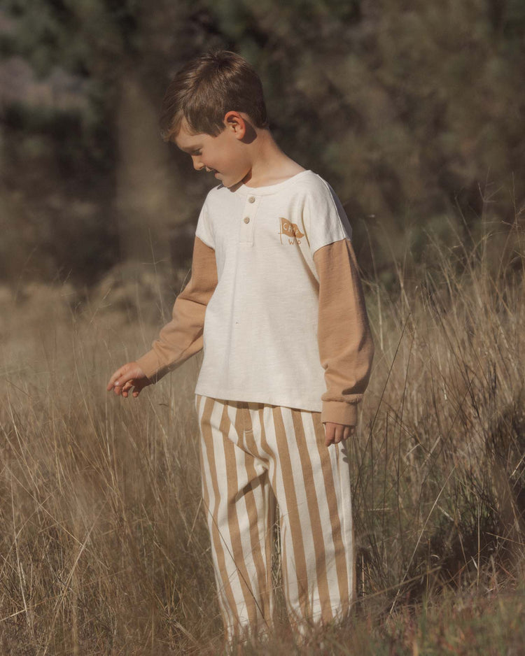 A young boy stands in tall grass outdoors, wearing the rylee + cru baggy pant in fawn pinstripe with an elastic waistband and a long-sleeve cream and tan shirt, looking down as he gently touches the grass.
