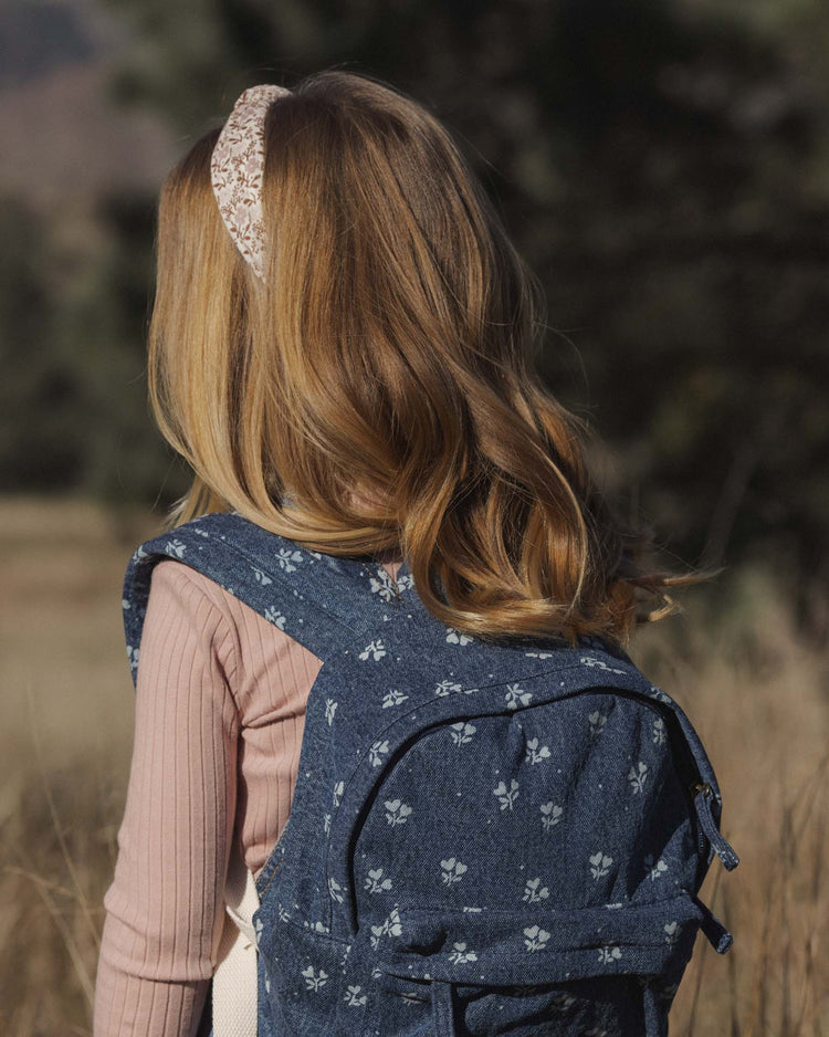 A young girl with long, wavy blonde hair, dressed in a pink headband and pink long-sleeve top, stands on a grassy outdoor field with blurred trees behind her. She carries the rylee + cru mini backpack in heart ditsy.