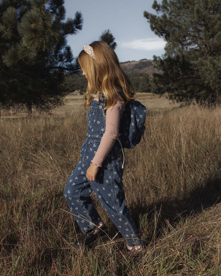 A young girl with long hair, dressed in indigo denim overalls featuring a charming heart ditsy print and a pink long-sleeve shirt, walks through tall grass in a field backed by pine trees. She carries the rylee + cru mini backpack in heart ditsy and wears a headband.