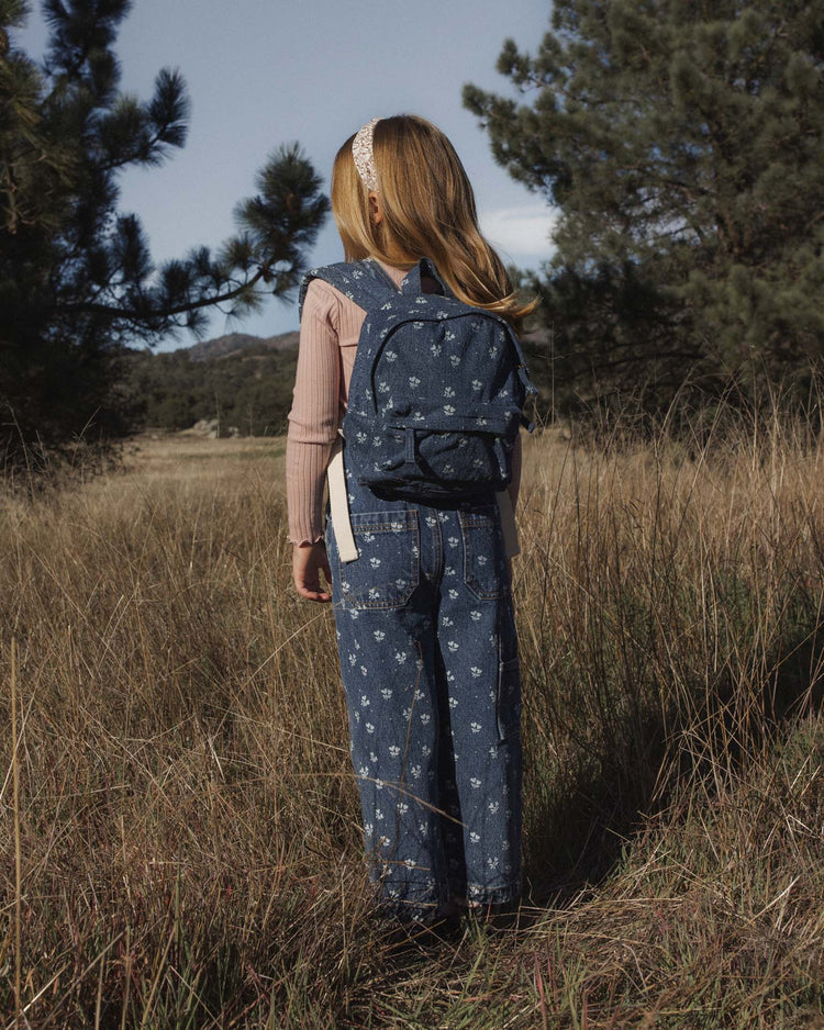 A young girl with long hair stands in a grassy field, facing away. She wears a headband, a pink long-sleeve shirt, indigo denim overalls with a heart ditsy print, and carries the rylee + cru mini backpack in heart ditsy on her back. Pine trees and hills are in the background.