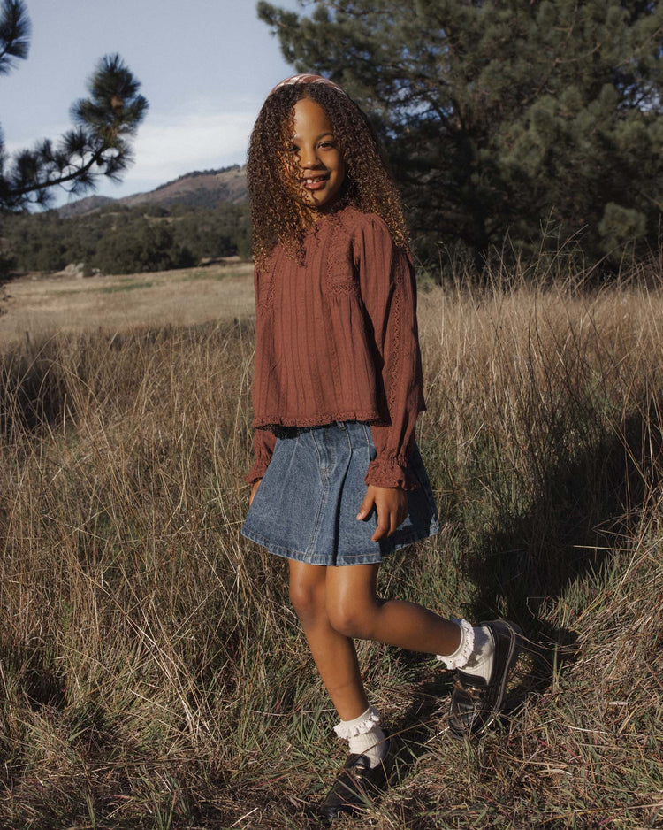 A young girl with curly hair smiles while standing in a grassy field, wearing the cecile blouse in mulberry by rylee + cru, paired with a denim skirt, white socks, and black shoes. Trees and hills create a scenic background.