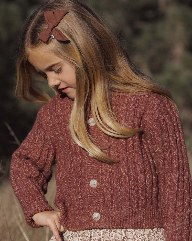 A young girl with long blonde hair and a brown bow looks down, wrapped in a cozy layer—a rylee + cru cheyenne cardigan in mulberry, featuring large marble buttons—over a floral dress, as she stands outdoors against a softly blurred natural background.