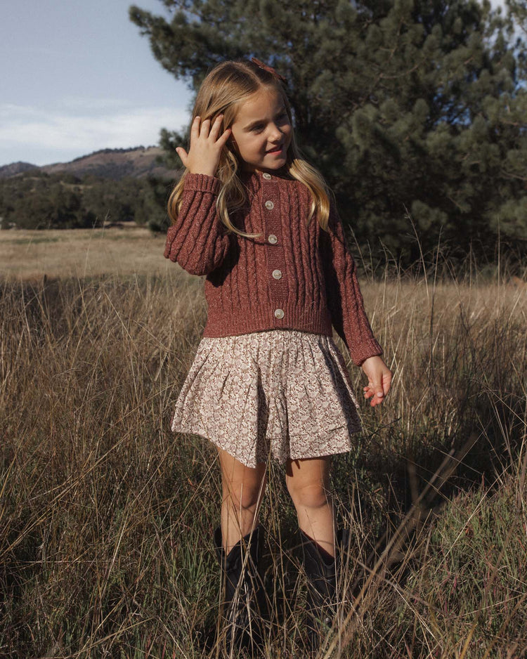 A young girl stands in a grassy field, framed by trees and hills, wearing the rylee + cru cheyenne cardigan in mulberry—a cozy knit layer with marble buttons—paired with a light-patterned skirt and black boots.
