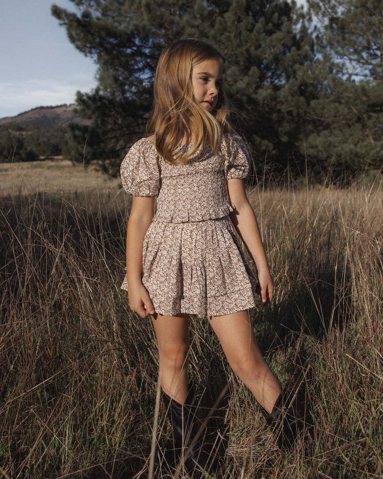 A young girl stands in a grassy field wearing the rylee + cru tiered mini skirt in primrose, styled with black boots. The 100% cotton skirt features tiers and is designed for comfort and ease of movement. Trees and mountains are visible in the background under a partly cloudy sky.