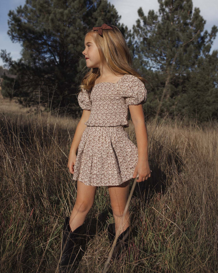 A young girl wearing the rylee + cru tiered mini skirt in primrose stands in tall grass, looking to the side, paired with brown boots and a floral top; trees and a cloudy sky fill the background.